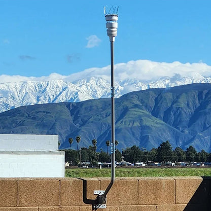 White Ecowitt device on a long pole secured to a wall with a bracket against a mountainous landscape under a blue sky | Lykalyte